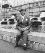 Man in a suit and hat sitting on a fountain ledge in front of a stone building