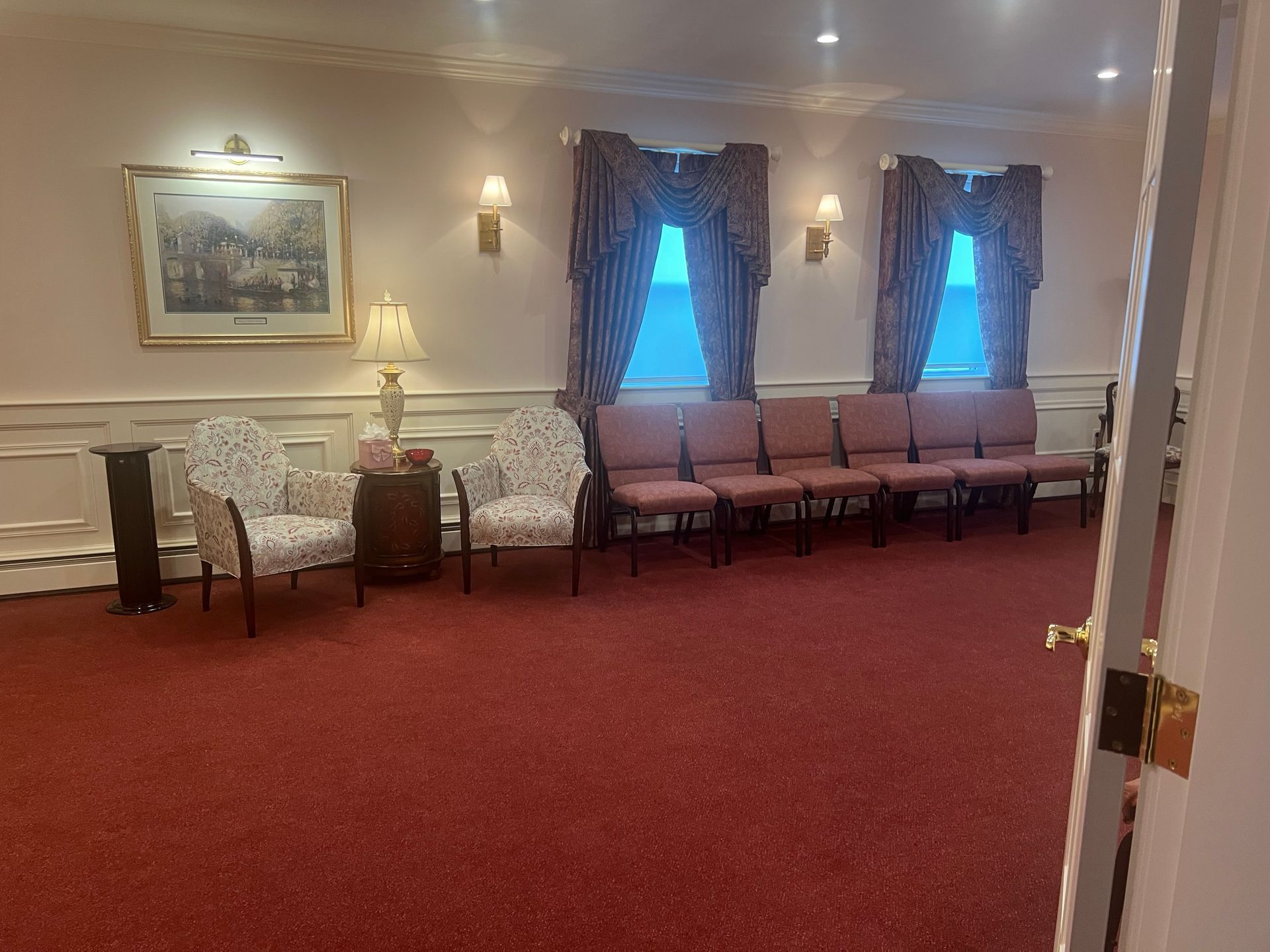 Elegant empty waiting room with red carpet, floral sofa, pink chairs, and curtained windows.