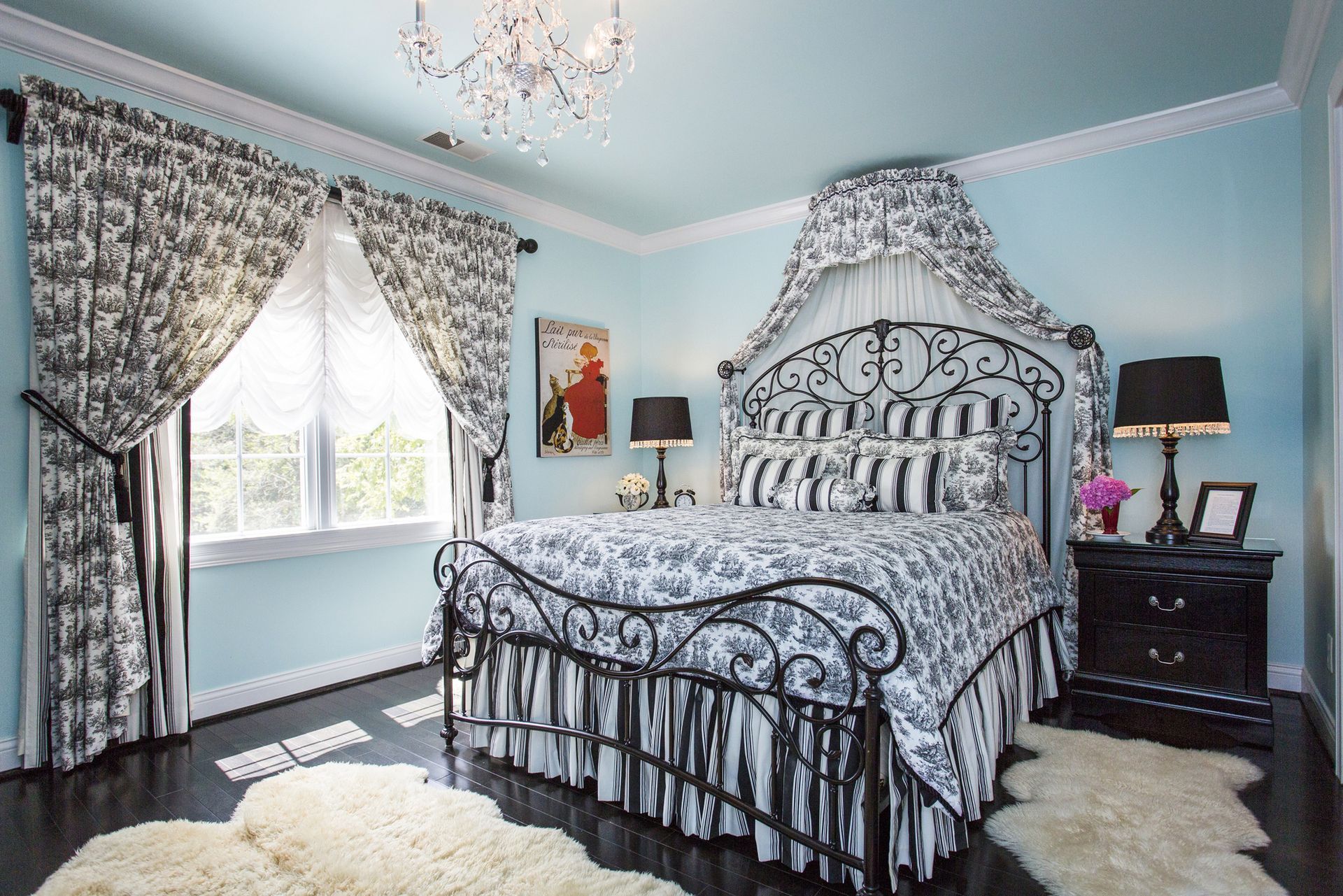 Bedroom with light blue walls, dark wood floor, ornate black bed frame and patterned bedding, curtains.