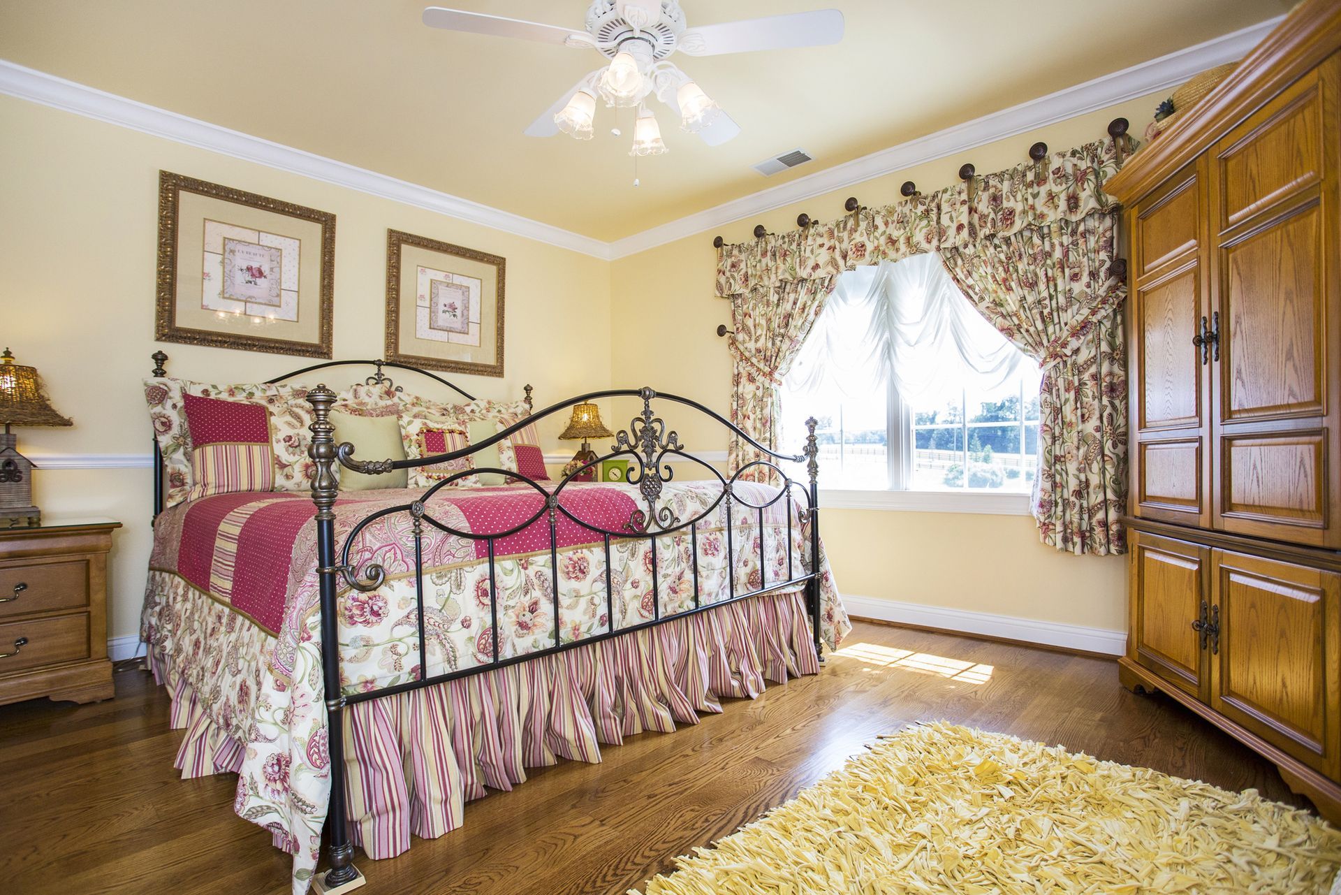Bedroom with floral bedspread, iron bed frame, wooden armoire, and window with patterned curtains.