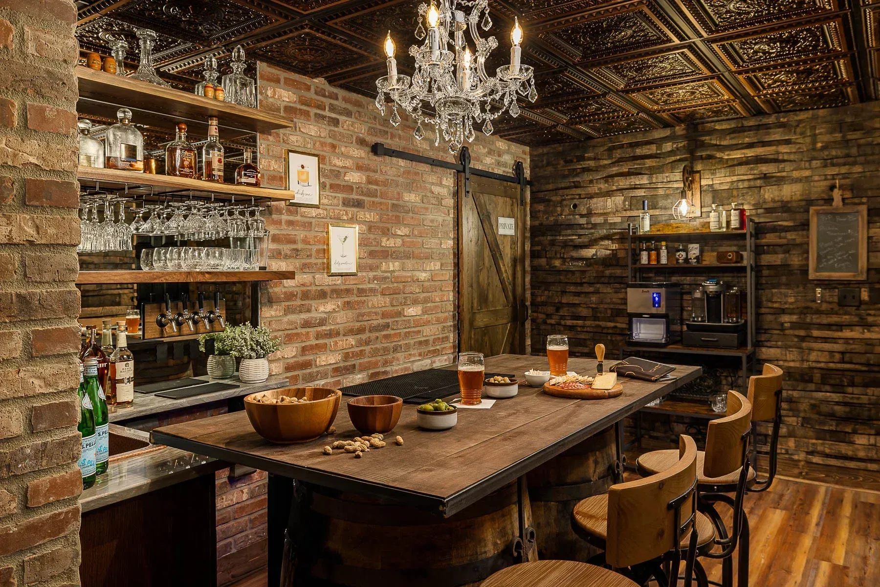 A home bar with brick walls, a chandelier, and a wooden bar top with snacks, drinks, and bar stools.
