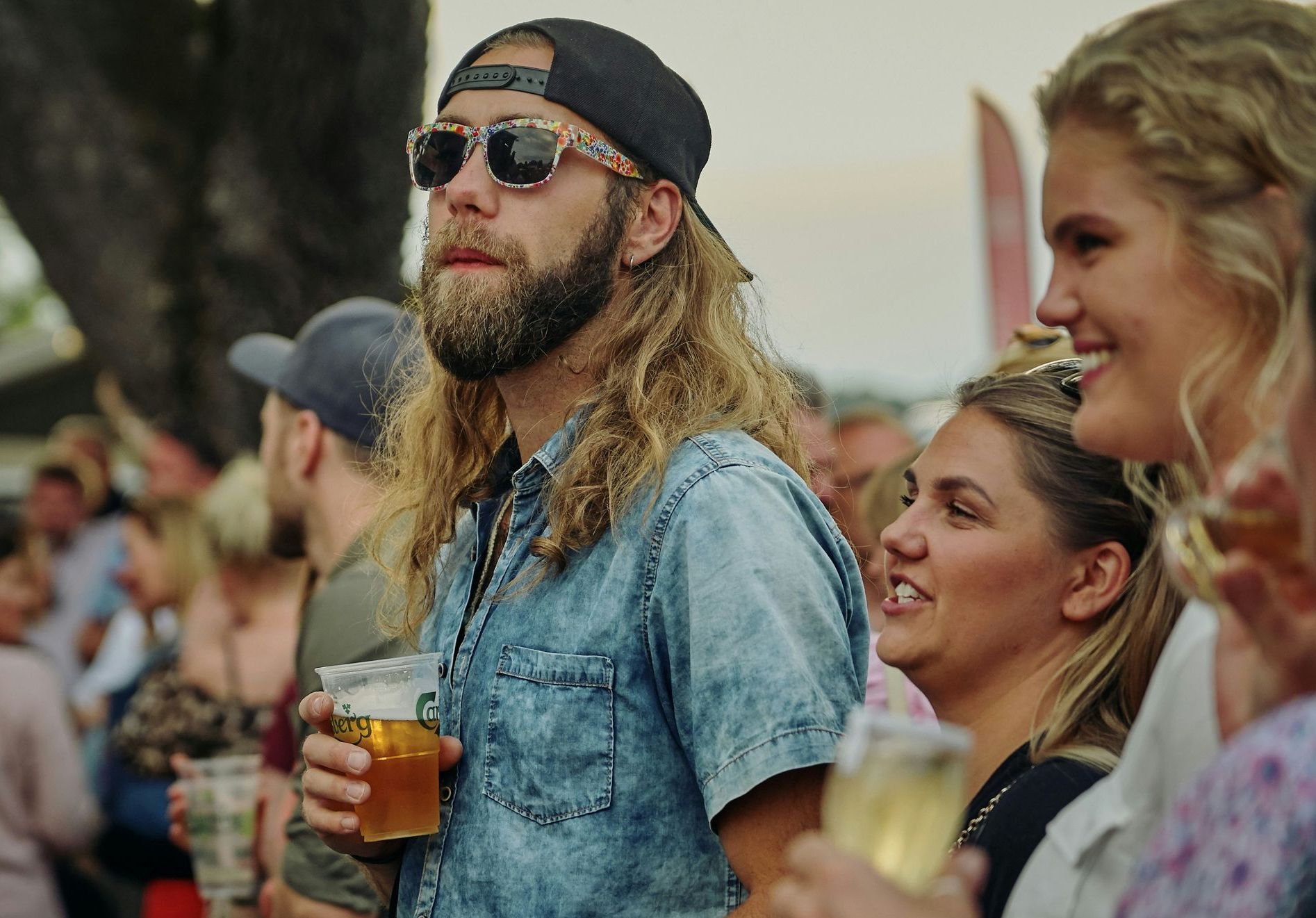 A man with a beard is holding a glass of beer in a crowd of people.
