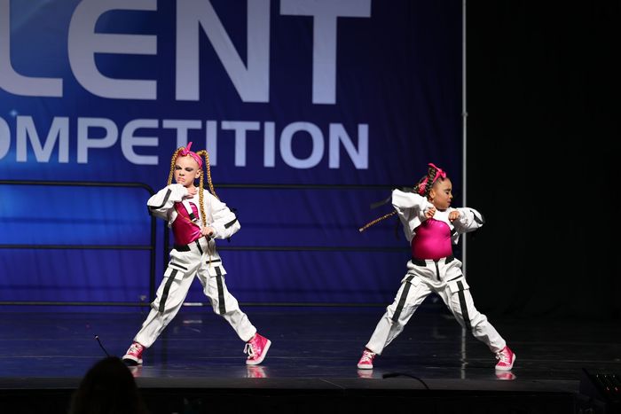 Two dancers on stage, wearing white and pink outfits, performing at a talent competition.