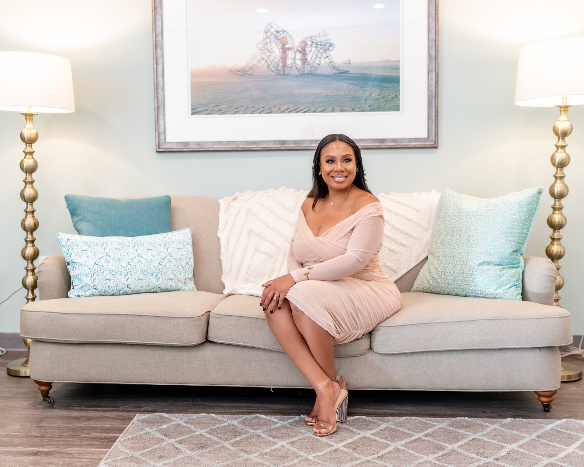 Woman seated on sofa, smiling. Soft beige dress, blue cushions, gold lamps, art on wall.