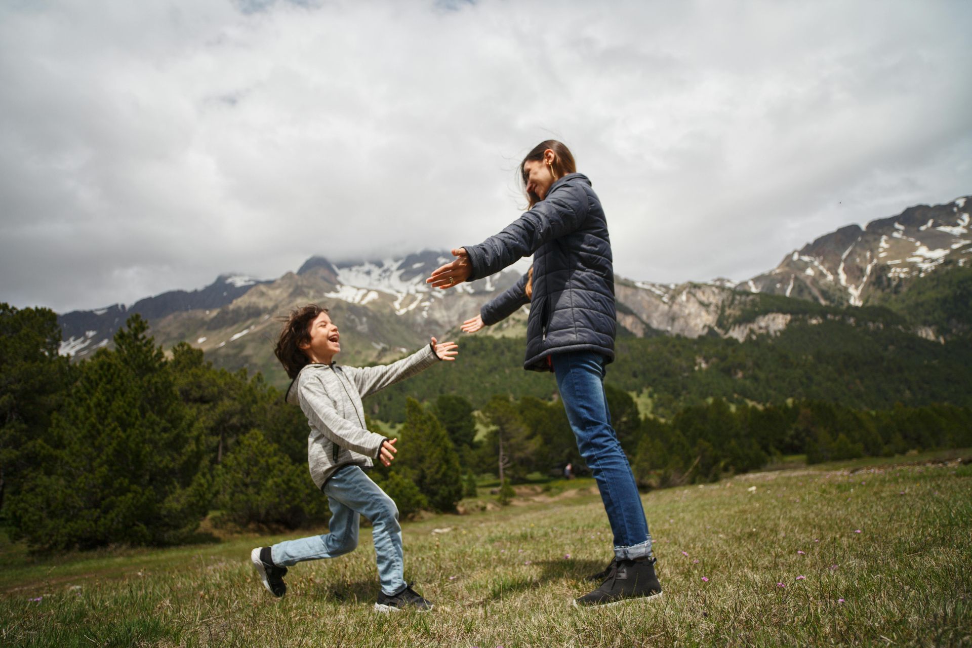 Person about to hug another in a mountain meadow, arms outstretched. Green grass, pine trees, snow-capped peaks in background.