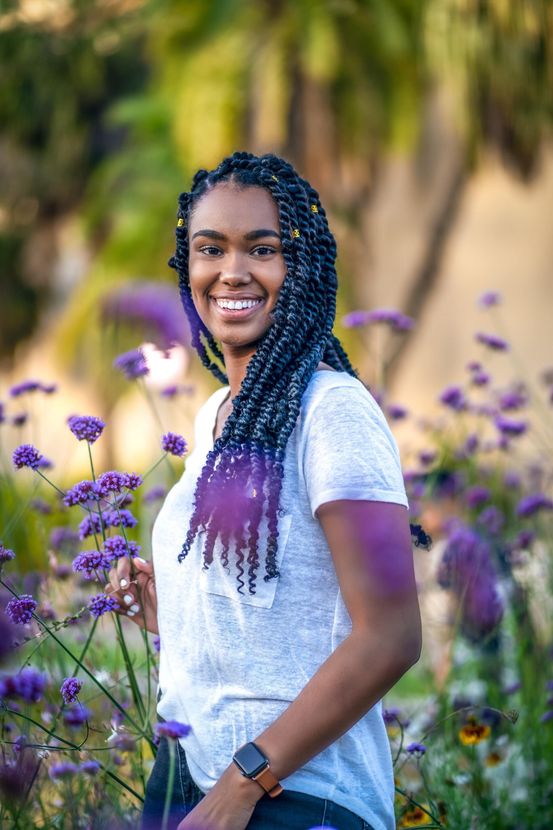 Woman smiling, holding purple flower, braided hair, in a garden of purple flowers, wearing a white shirt and watch.