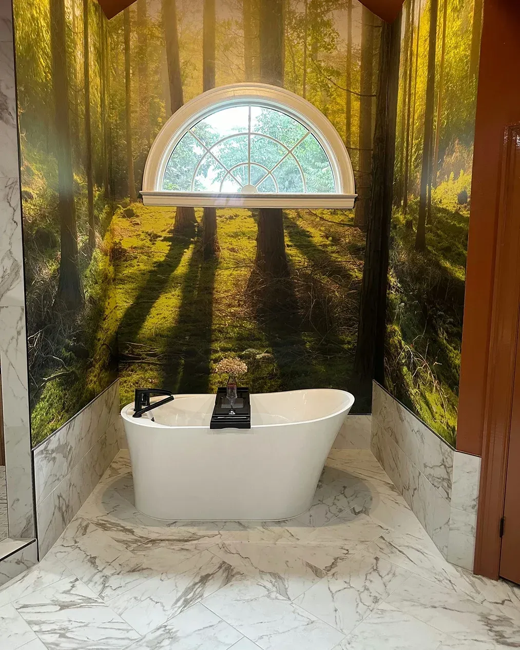 Woodland-themed bathroom with a white bathtub beneath an arched window, marble floor, and a forest mural