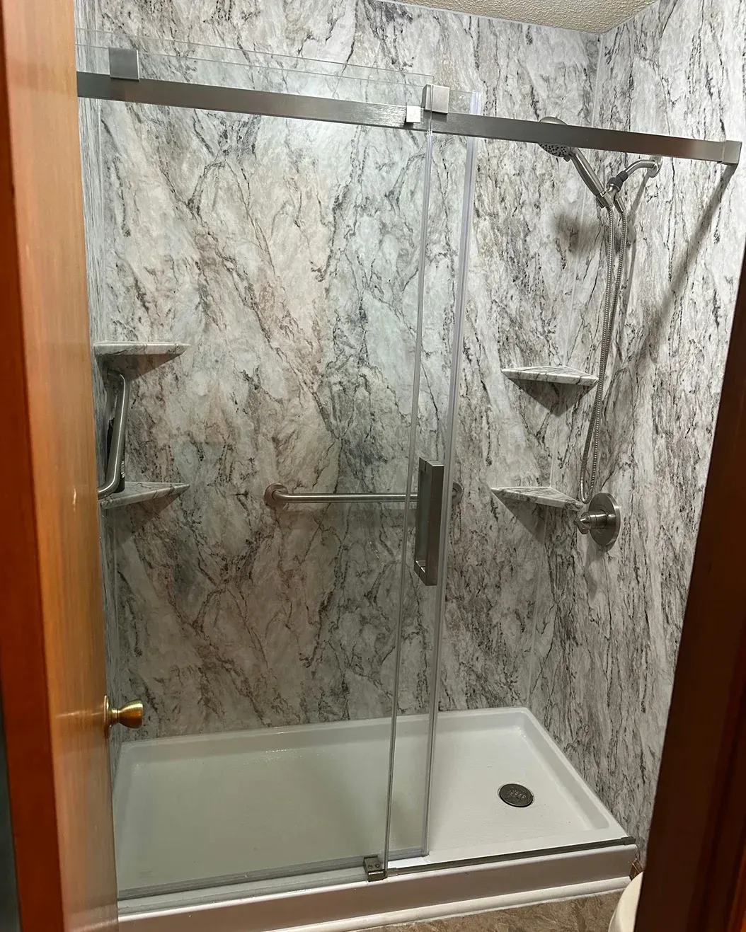 Glass shower with marble-pattern walls and white base, seen through a partially open door.