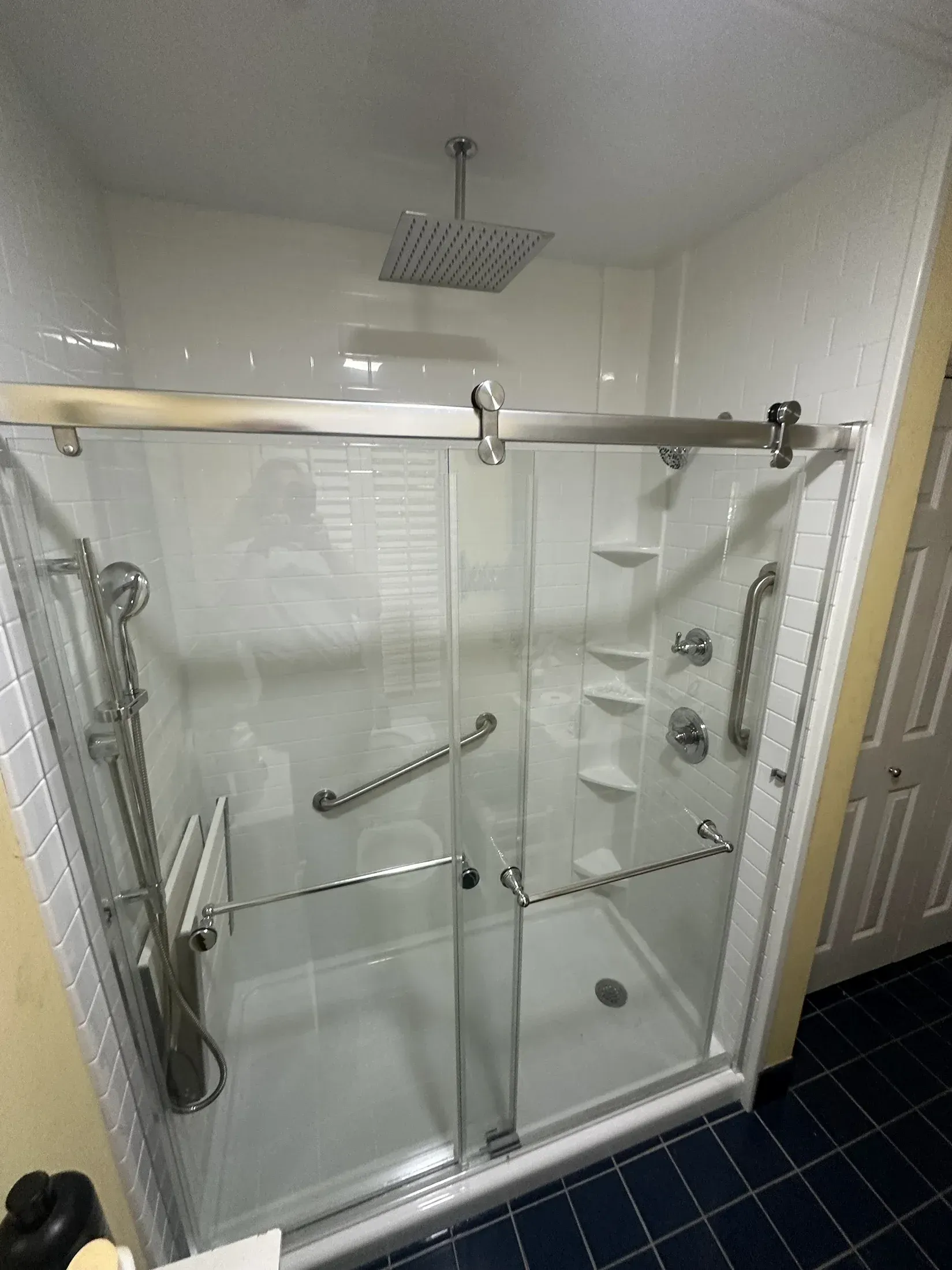 Accessible walk-in shower with white tile walls, glass doors, grab bars, and fold-down seat.