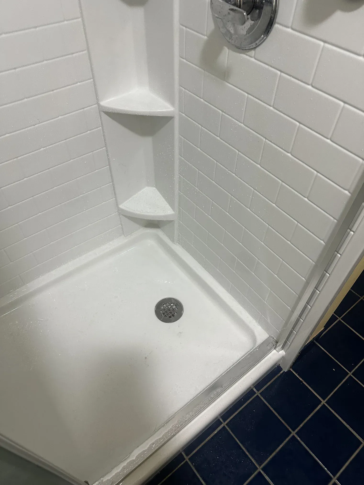 White tiled shower stall with a drain, corner shelves, and dark blue floor tiles outside the threshold