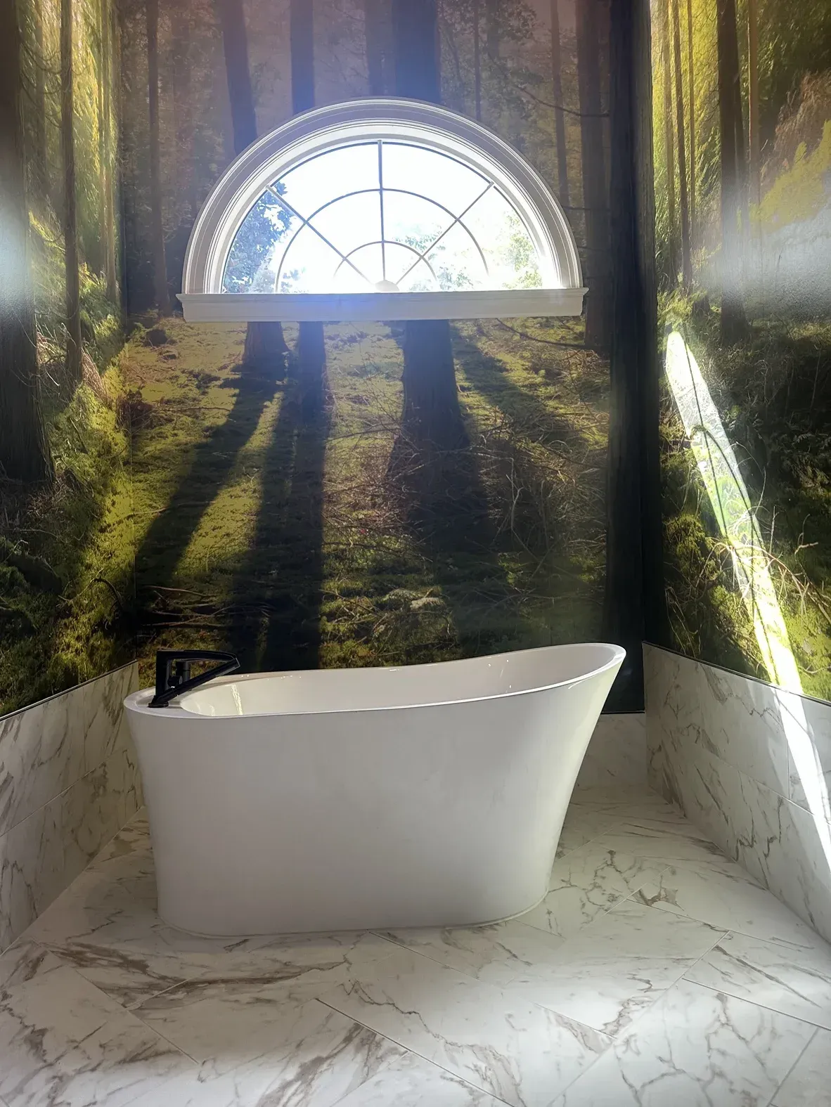 White freestanding bathtub beneath an arched window, in a bright, sunlit bathroom with forest mural walls.