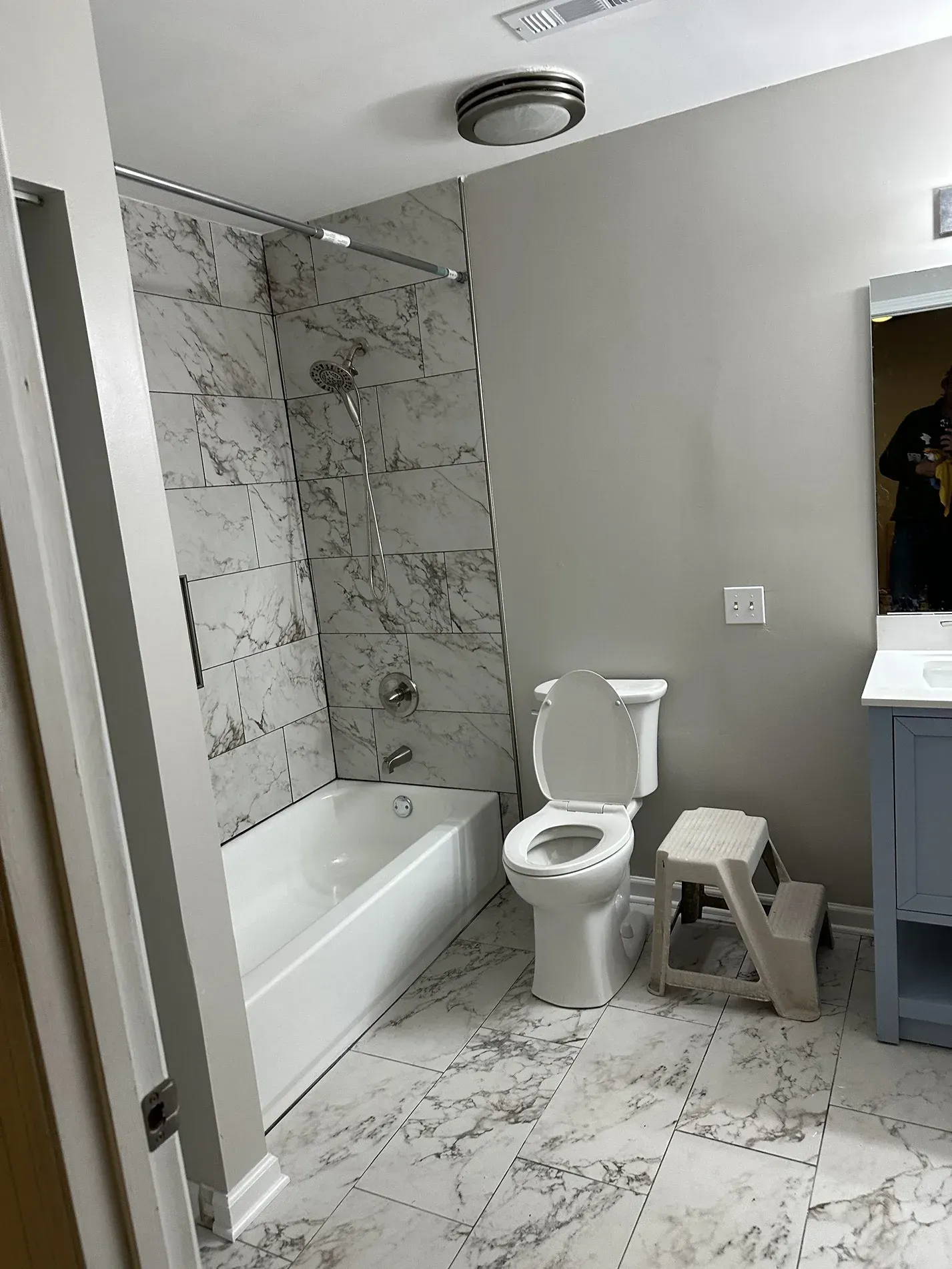 Small gray bathroom with marble tiles, bathtub, toilet, stool, and vanity mirror.
