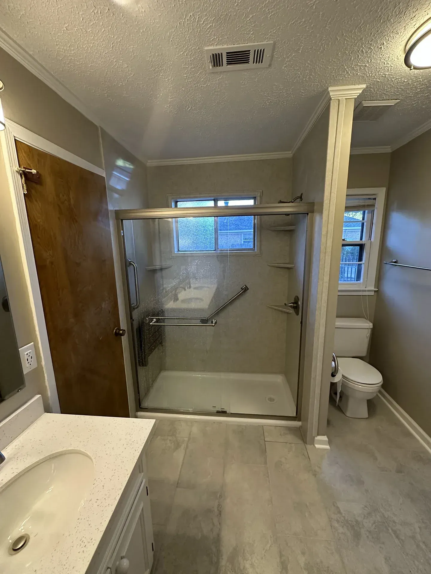 Small bathroom with a walk-in shower, toilet, sink, and grab bars under a window.