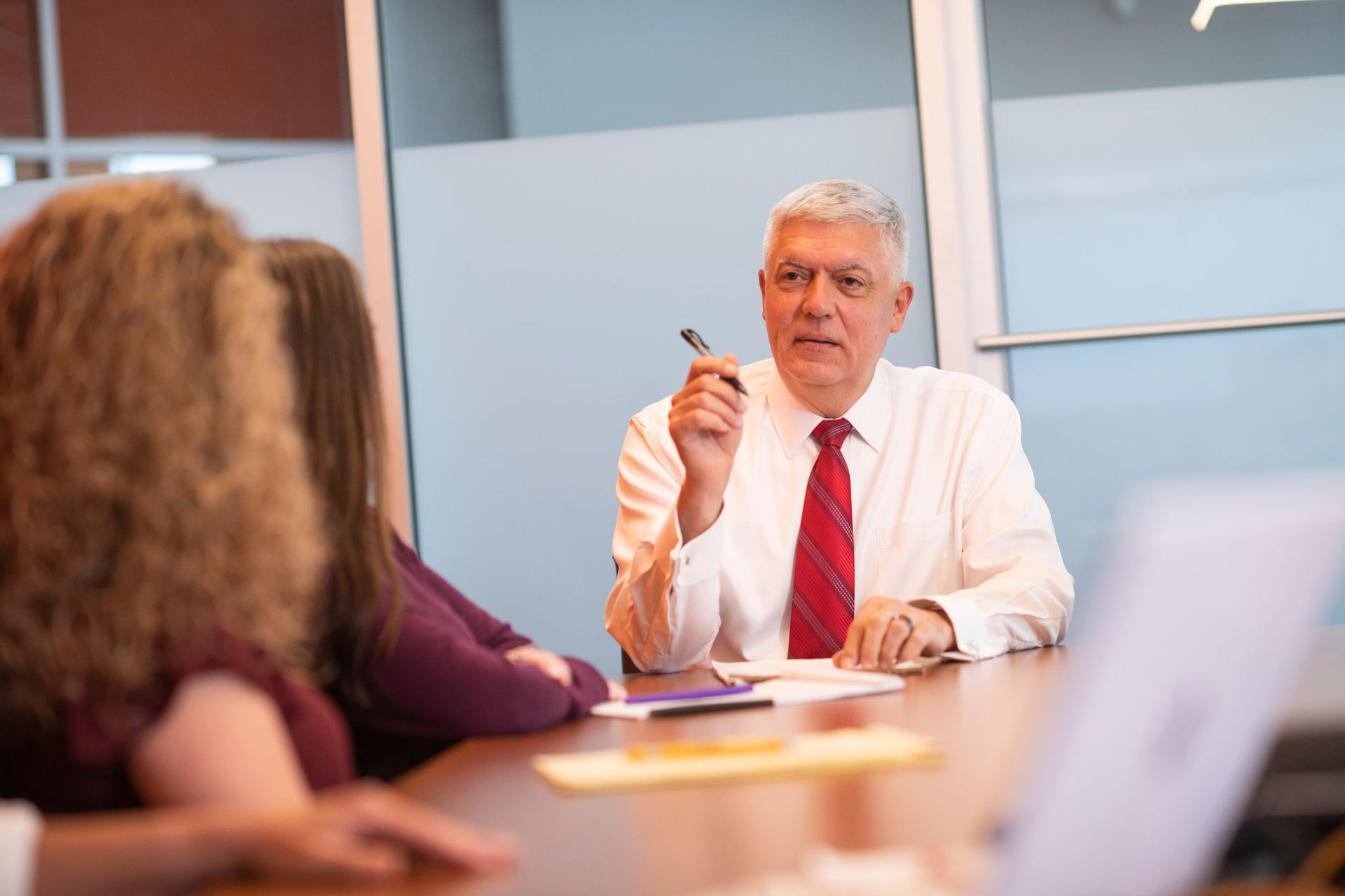 Business meeting in a conference room, with a man in a white shirt and red tie speaking at a table.