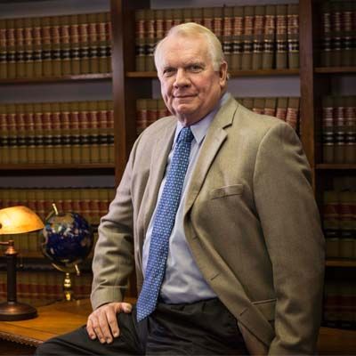 Man in tan suit seated in an office with law books and a globe behind him