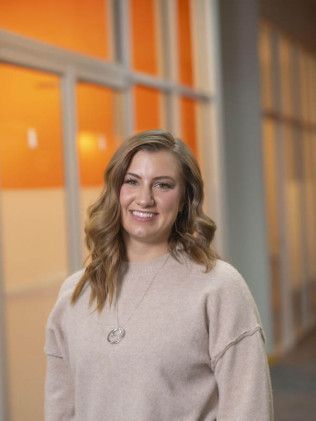 Woman smiling in a beige sweater, standing in a hallway with orange and gray walls.