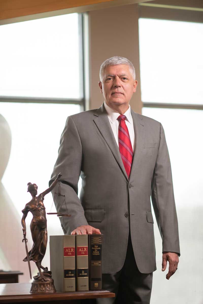Man in a gray suit with red tie stands beside a trophy and law books in an office.