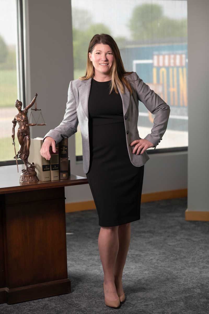 Woman in a gray blazer and black dress standing by an office desk and large window.