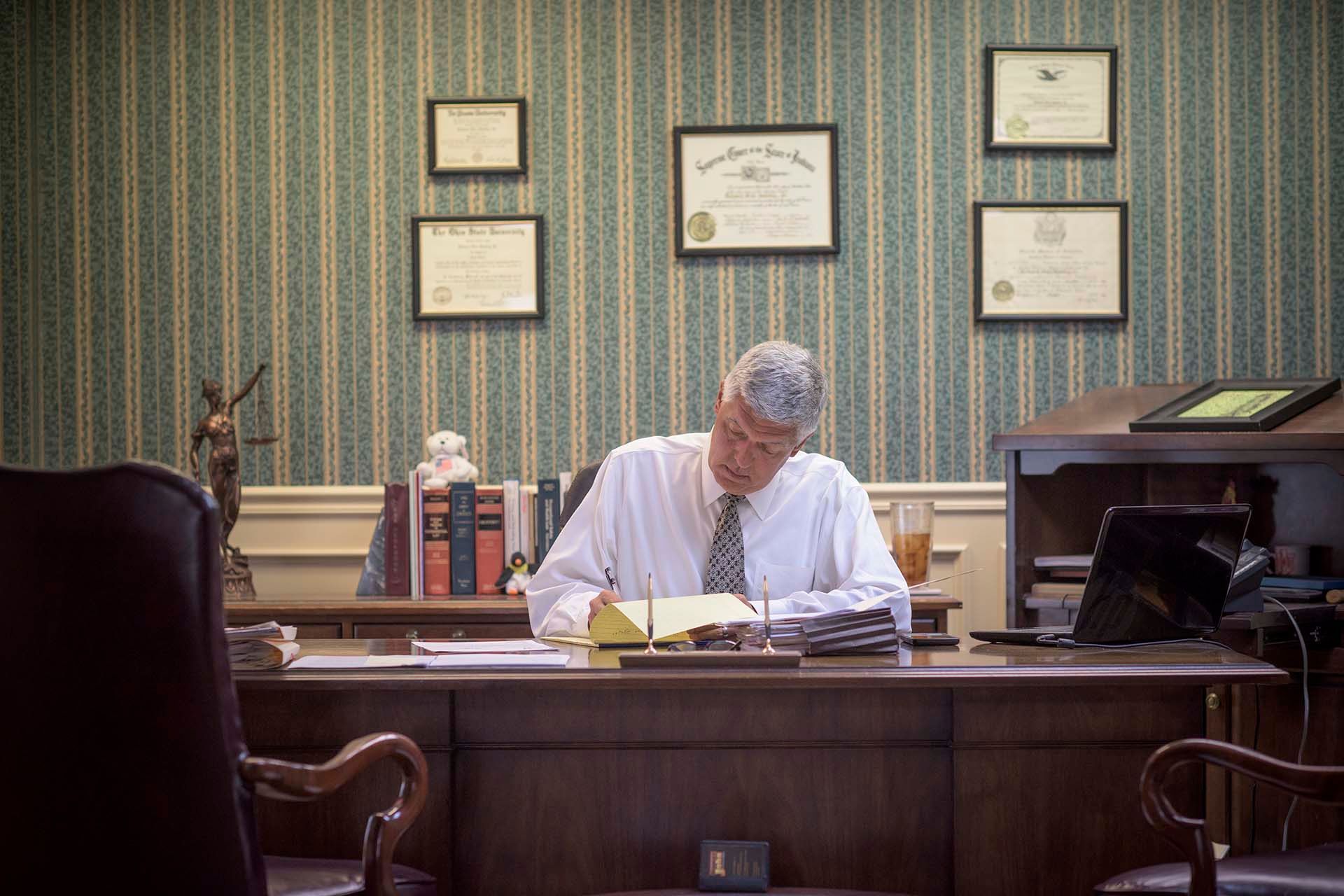 Man in a white shirt writing at a desk in an office with framed certificates on the wall