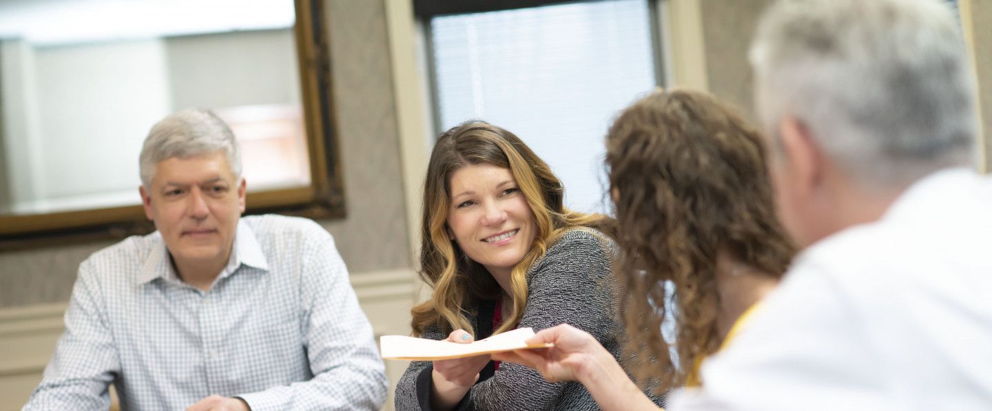People in a meeting passing a document across the table, smiling in a bright office