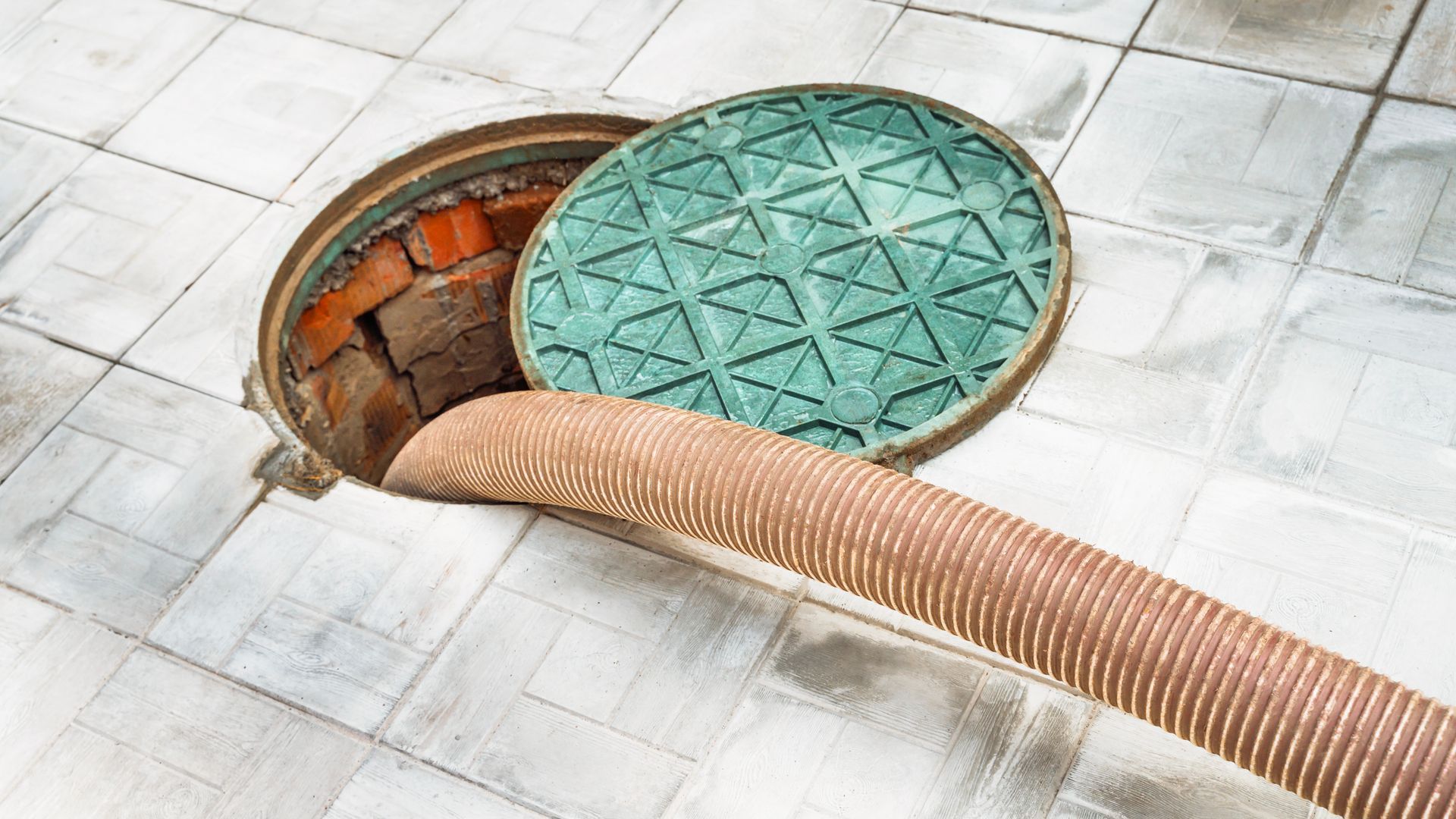 Open manhole with green cover partially lifted and beige hose inserted on tiled pavement