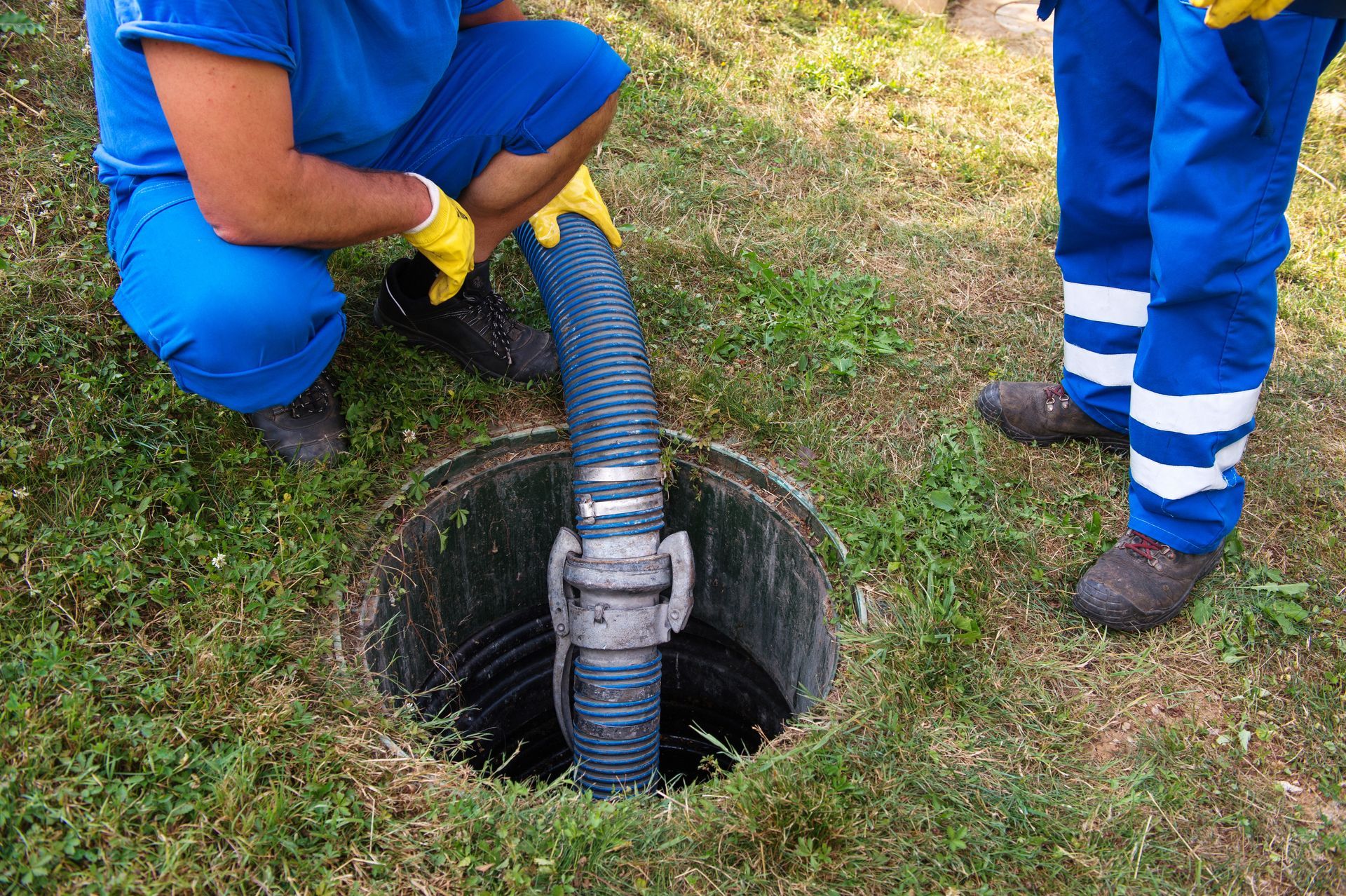 Two workers in blue uniforms use a hose to inspect a manhole in a grassy area.
