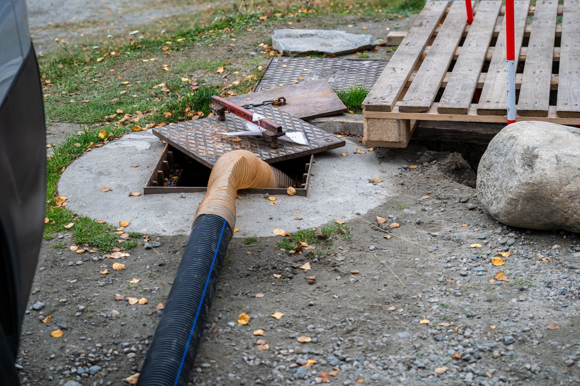Broken sidewalk with a fallen utility pipe, a metal grate, and a wooden pallet on gravel near a curb