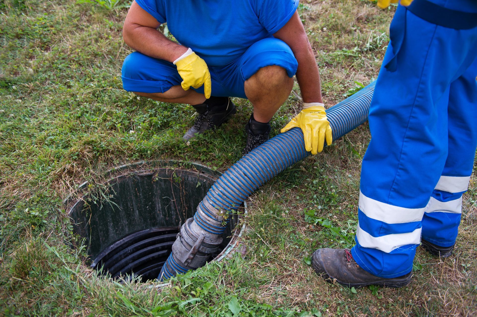 Two workers in blue uniforms use a hose to clean a roadside drain hole.