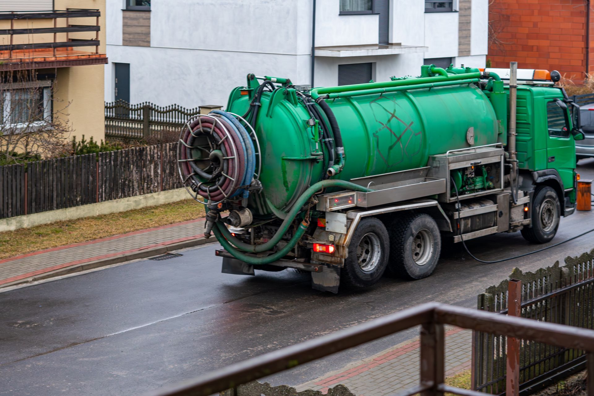 Green industrial vacuum truck on a wet residential street, viewed from the side.