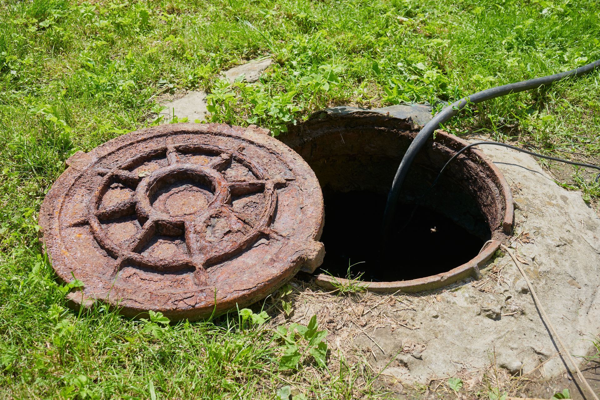 Open manhole with rusty lid beside a black hose on grass and concrete