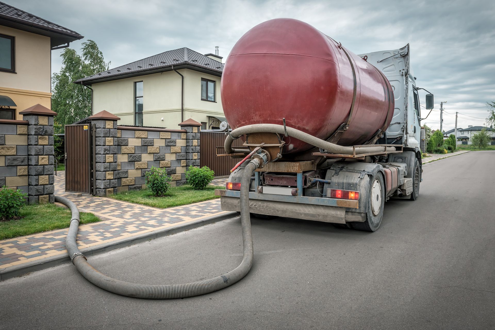 Red vacuum tanker hose stretched across a residential street near houses and a stone wall.