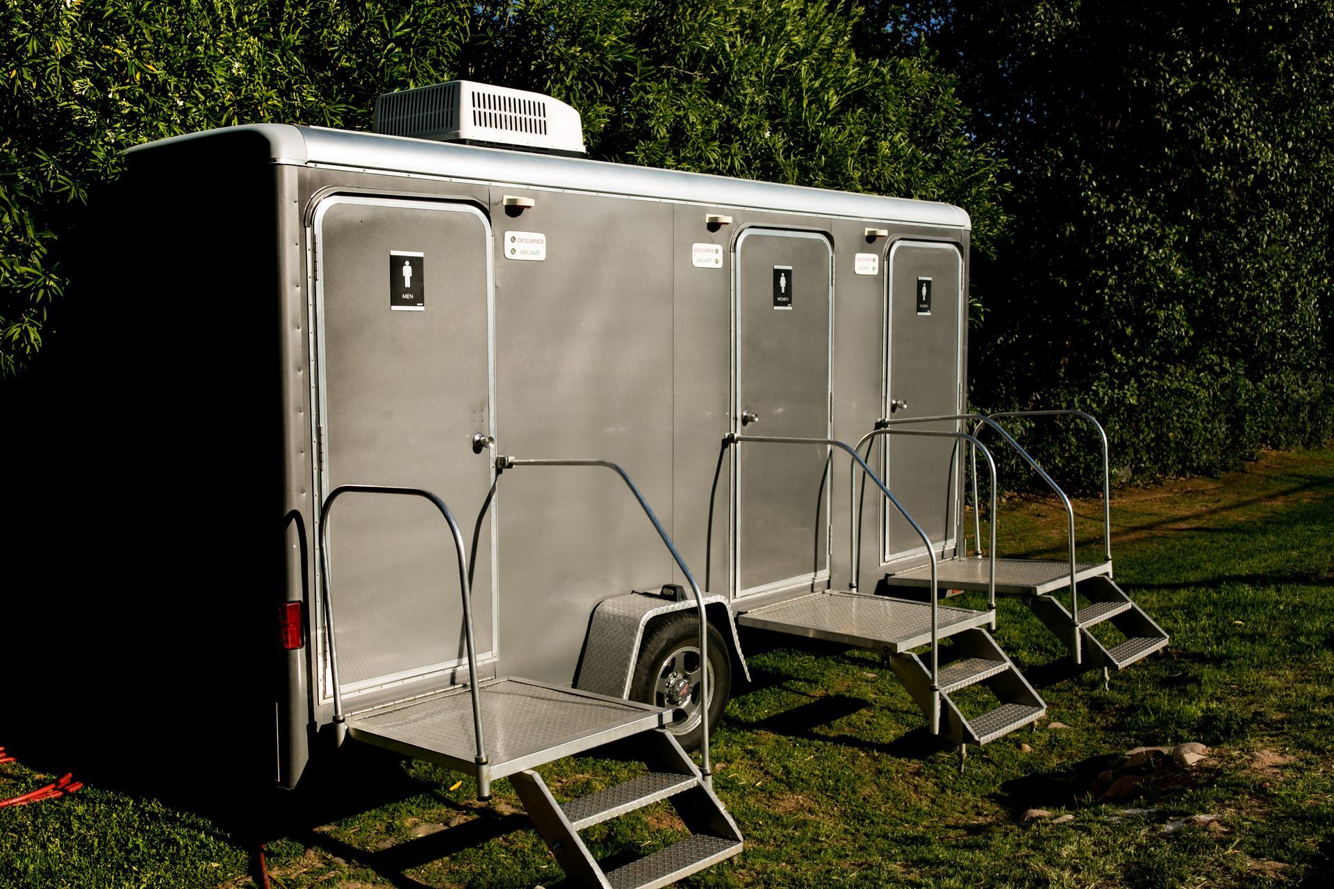 Silver portable toilet trailer with three doors and steps, parked on grass near trees.