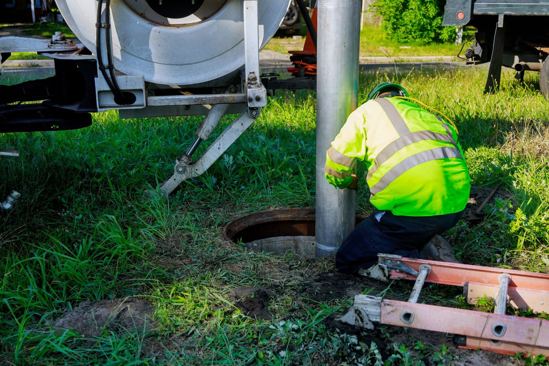 Worker in a neon safety jacket crouches beside an open manhole near heavy equipment in a grassy area.
