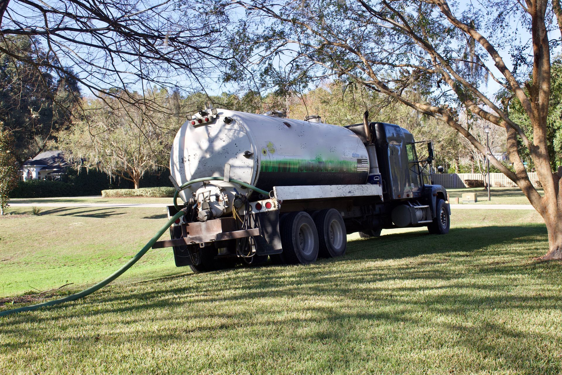 Vacuum truck parked on grass under trees, hose extended on a sunny day