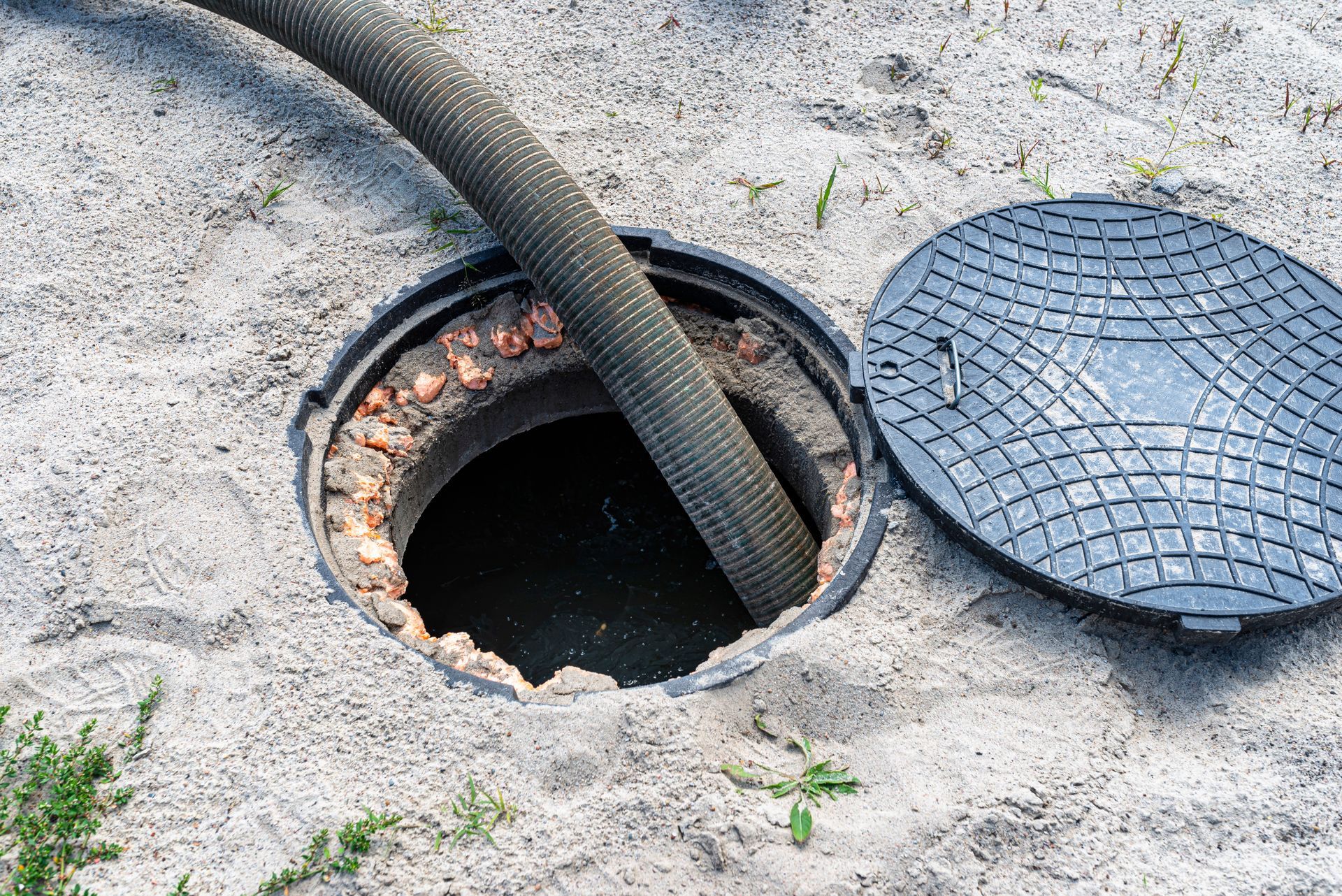 Open manhole with hose inserted, surrounded by sand, beside a metal cover.