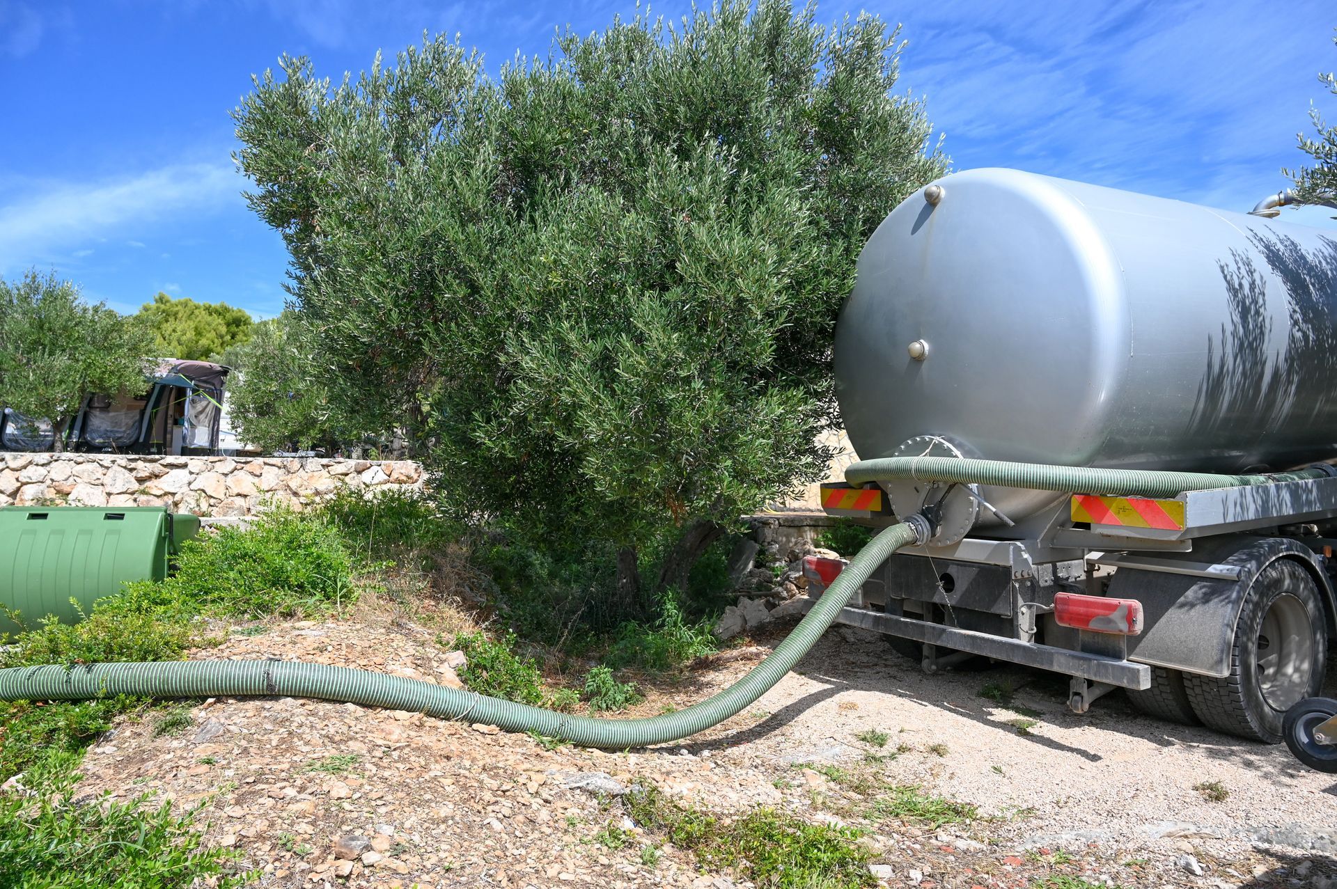 Silver water tanker truck pumping water near a leafy tree in a dry outdoor area