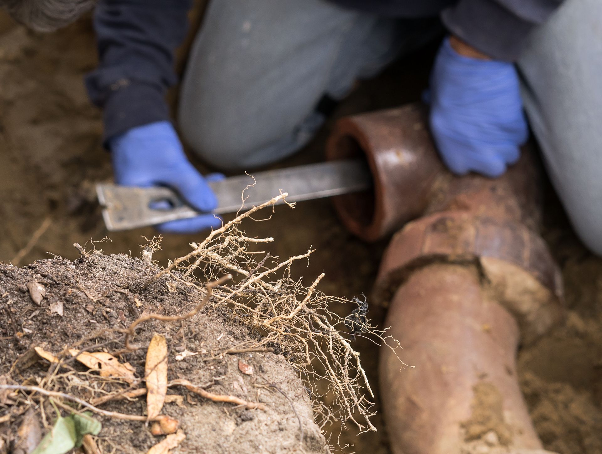 Gloved hands using a tool to inspect a rusted underground pipe in dirt