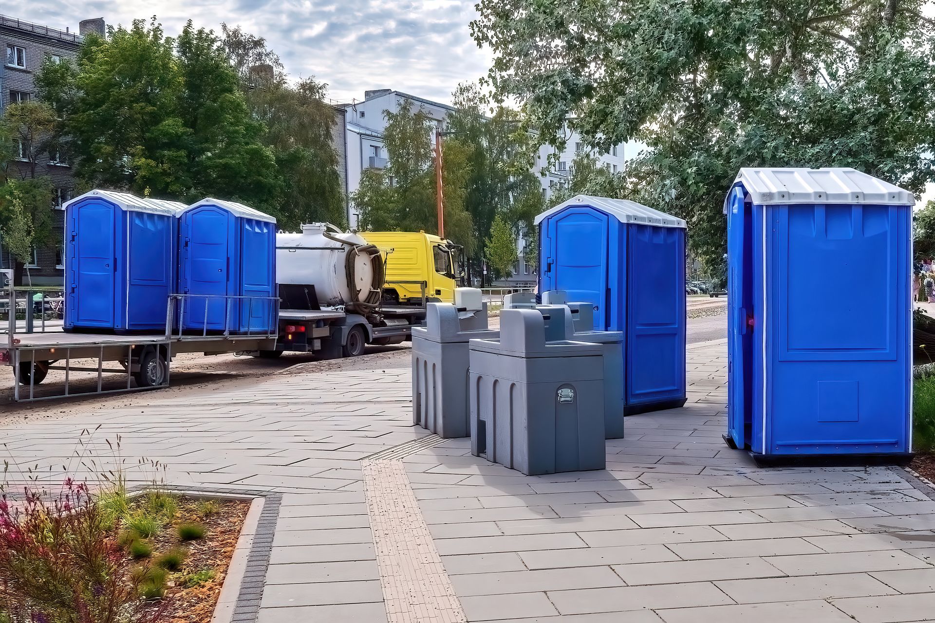 Blue portable toilets and gray barriers on a paved street near parked trucks and trees