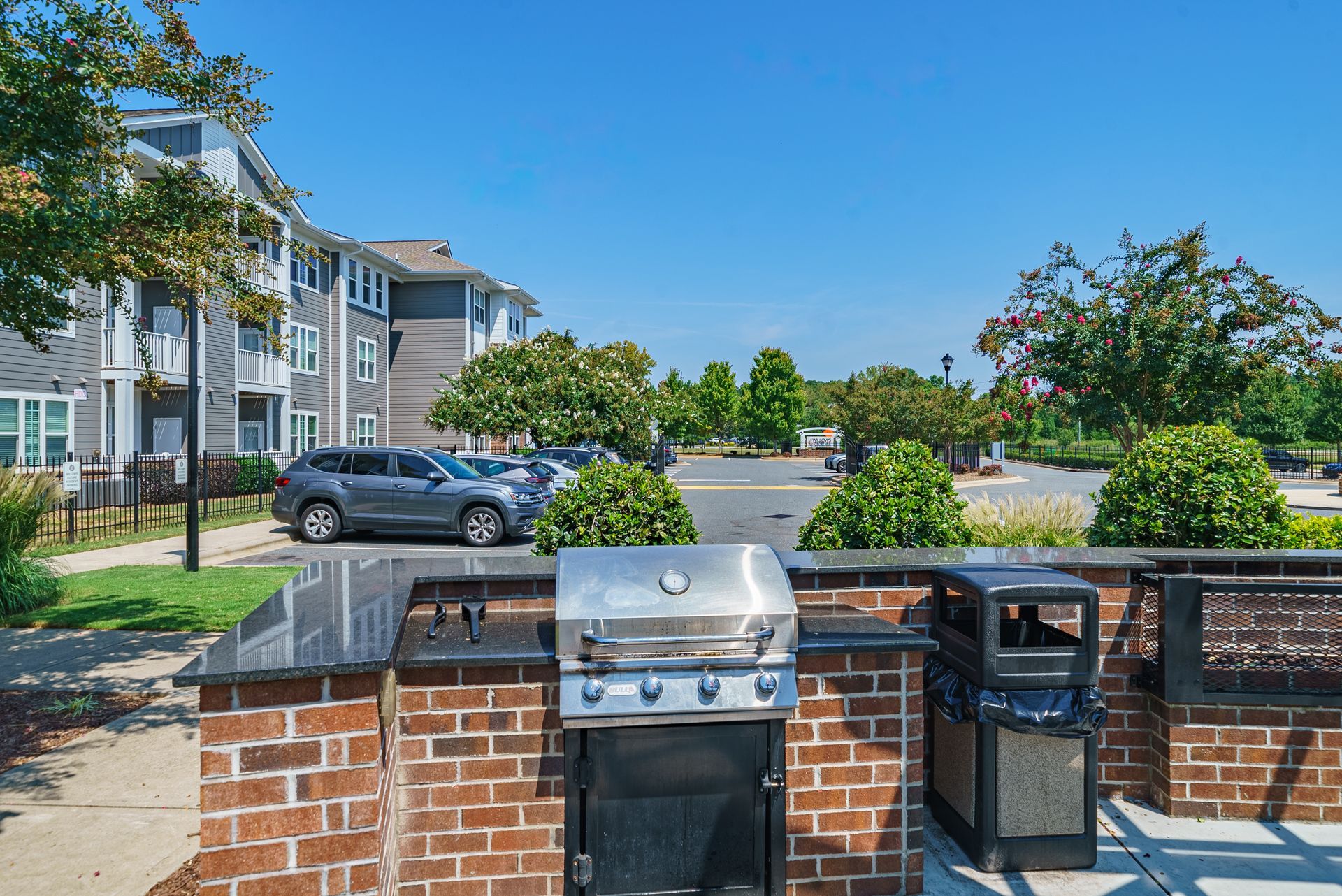 Apartment parking lot with brick grill station, black car, and apartment buildings under a clear blue sky at Willows at Fort Mill apartments for rent in SC.