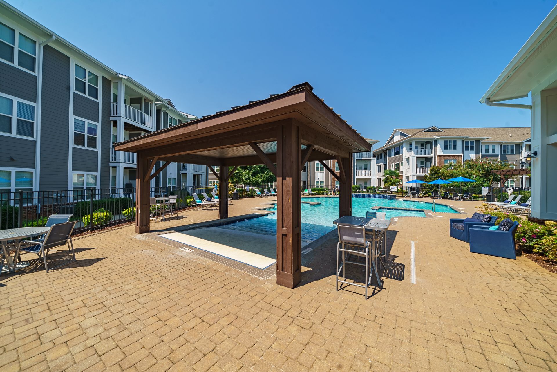 Resort pool area with covered cabana, blue lounge chairs, and brick patio under a clear sky at Willows at Fort Mill apartments for rent in SC.