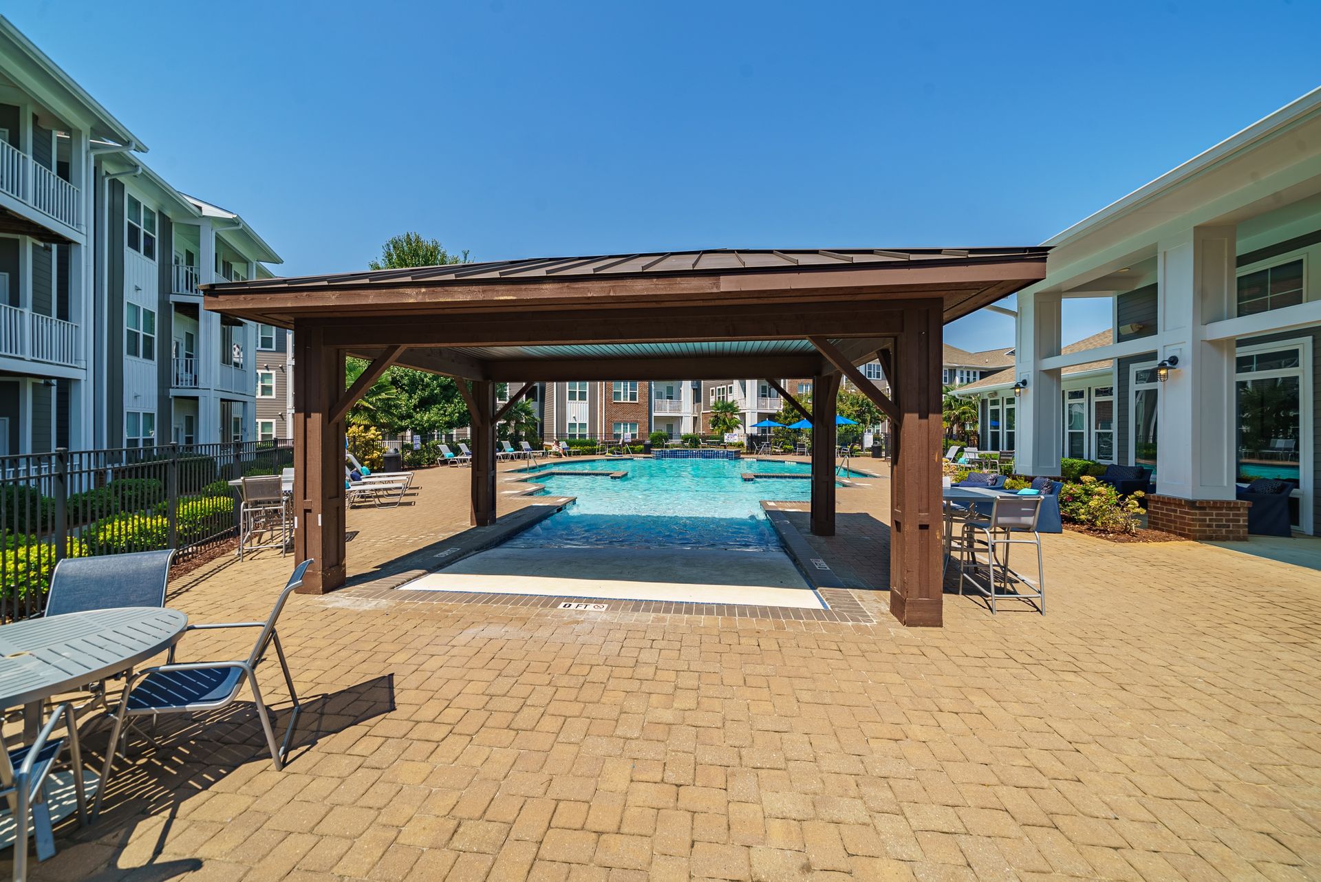 Resort pool deck with a wooden pergola, lounge chairs, and turquoise water between modern buildings at Willows at Fort Mill apartments for rent in SC.