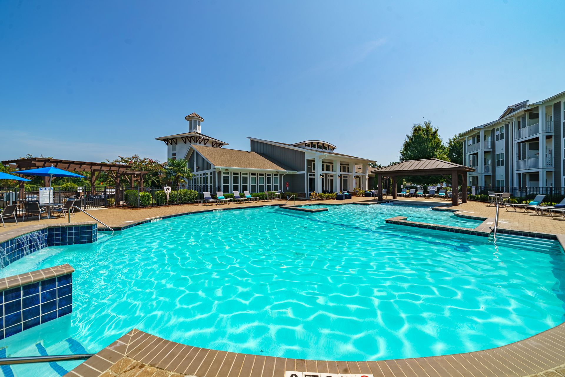 Outdoor resort swimming pool with turquoise water, lounge chairs, and modern buildings under a clear blue sky at Willows at Fort Mill apartments for rent in SC.