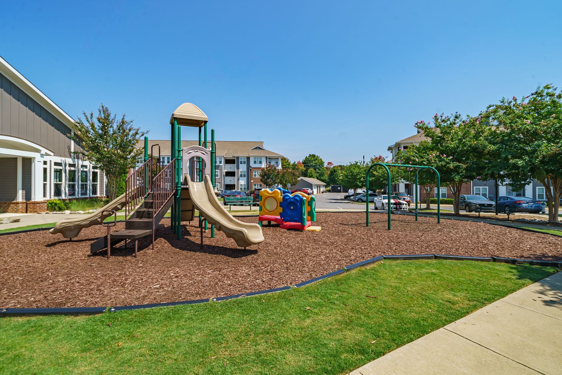 Playground with slides and a climbing tower in a sunny park beside a sidewalk and grassy lawn at Willows at Fort Mill apartments for rent in SC.