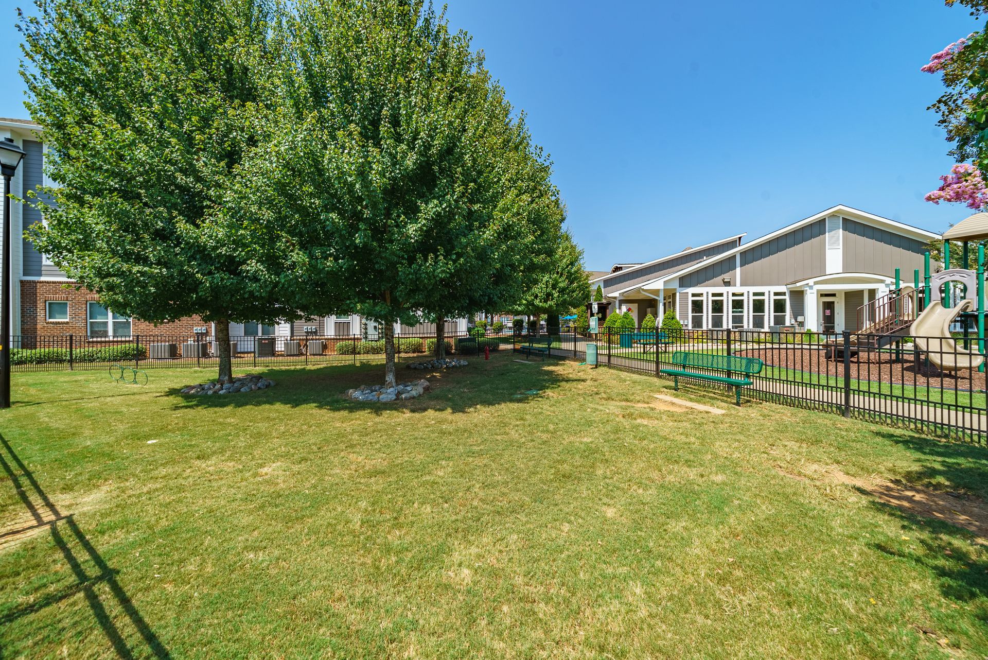 Sunny green park with trees, grass, and a low building beside a fenced walkway at Willows at Fort Mill apartments for rent in SC.