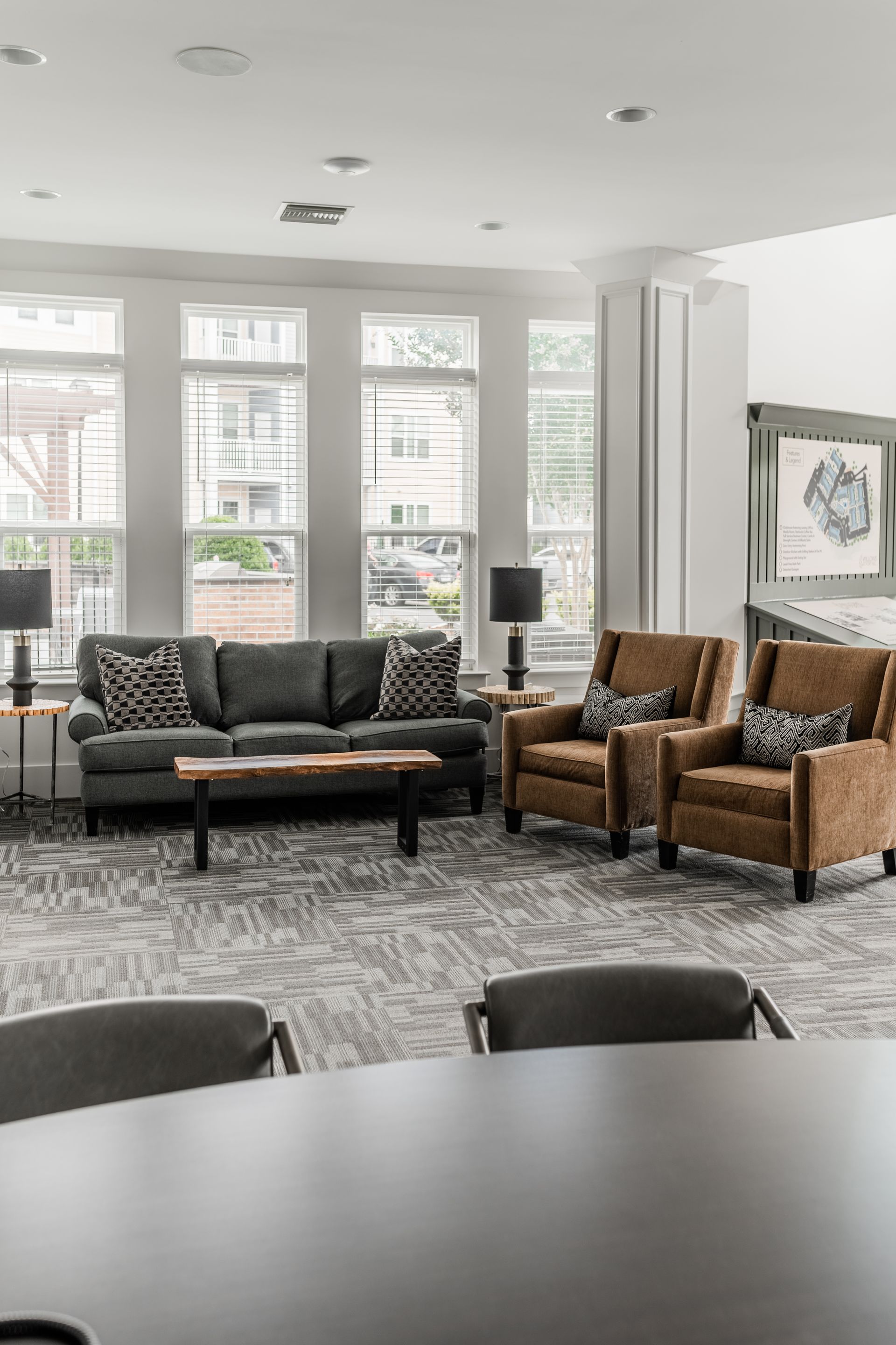 Bright living room with dark sofa, tan chairs, patterned rug, large windows, and dining table in foreground at Willows at Fort Mill apartments for rent in SC.