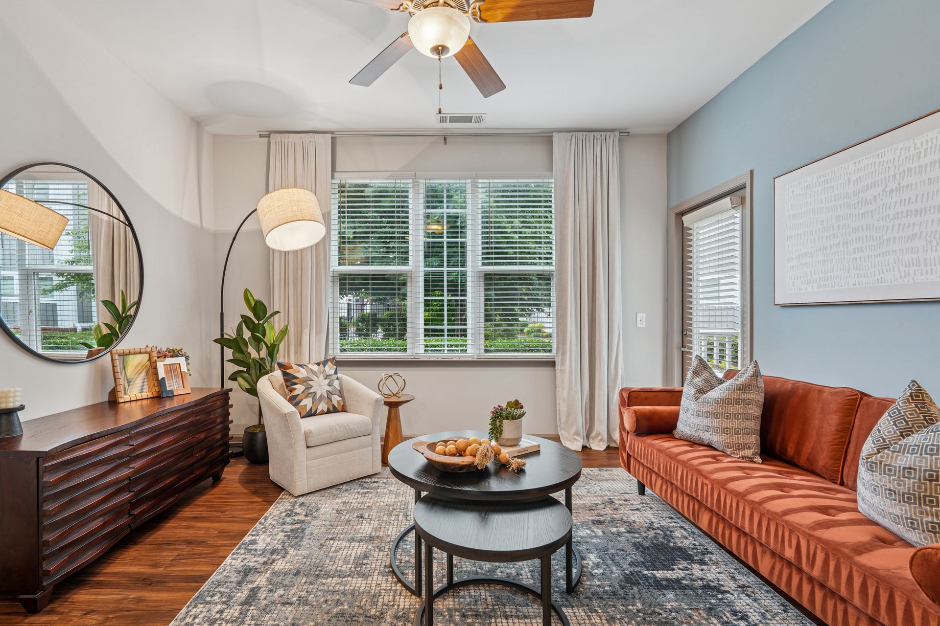 Bright living room with tan sofa, patterned chair, round coffee table, blue wall, and large window blinds at Willows at Fort Mill apartments for rent in SC.