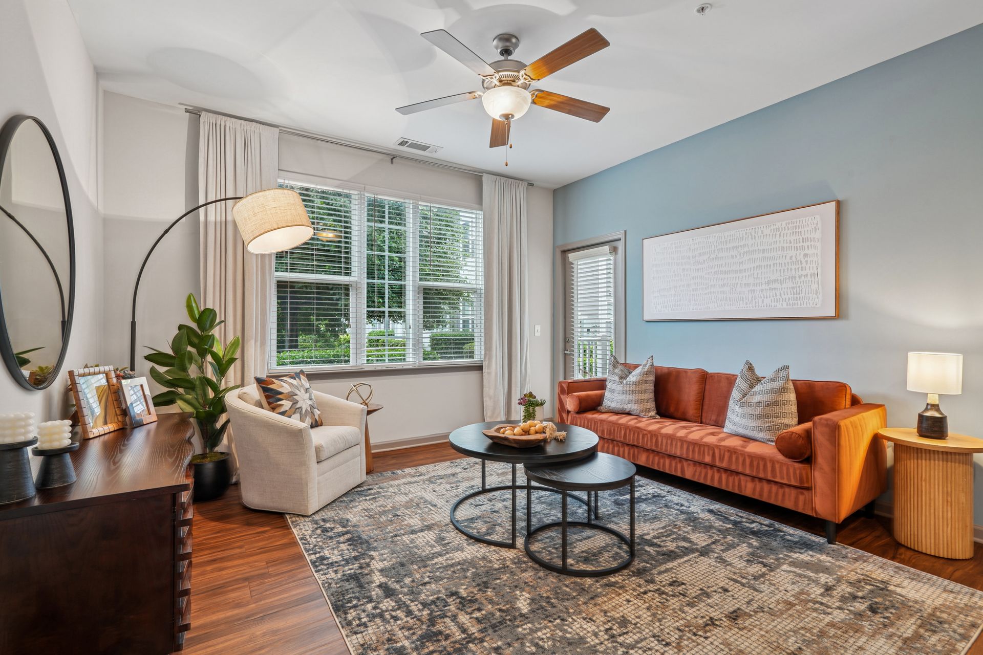 Bright living room with orange sofa, cream chair, large window, patterned rug, and ceiling fan at Willows at Fort Mill apartments for rent in SC.