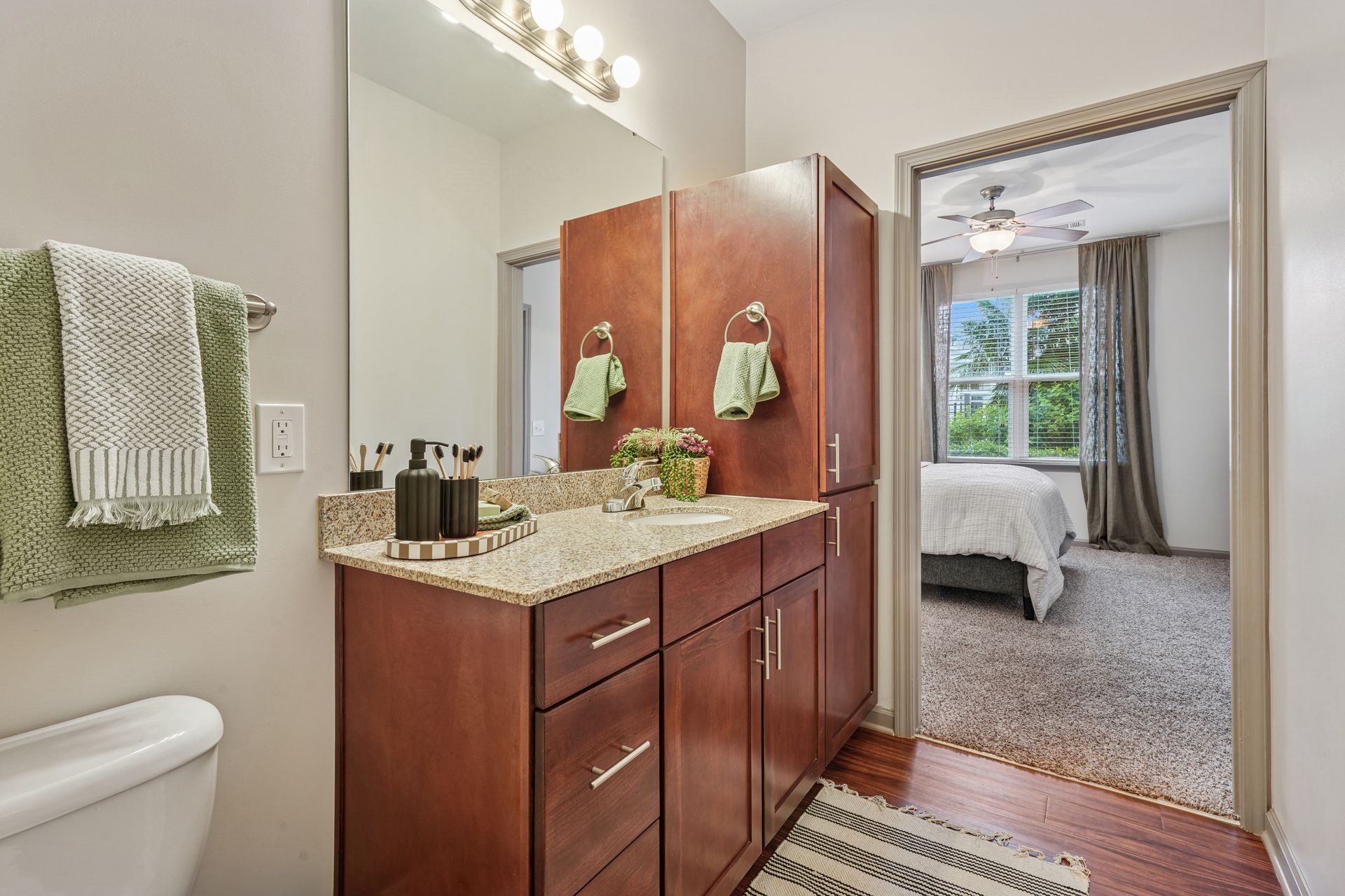 Bathroom with wood vanity, granite counter, and large mirror, viewed toward a bedroom with a bed and window at Willows at Fort Mill apartments for rent in SC.