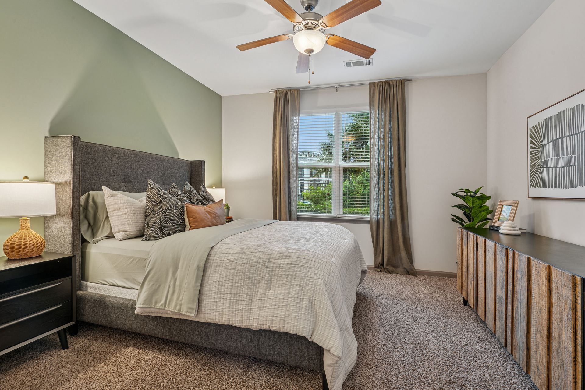 Bright bedroom with a gray bed, carpeted floor, window, ceiling fan, and striped dresser at Willows at Fort Mill apartments for rent in SC.