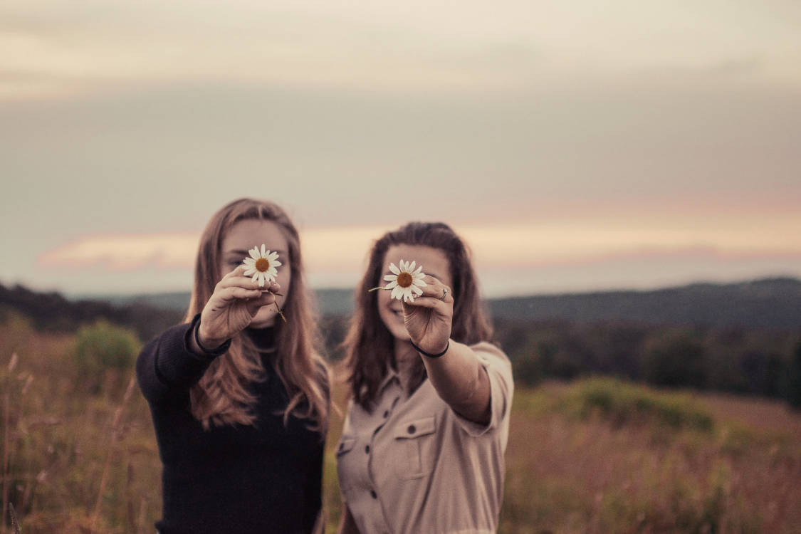 Two women are holding daisies in front of their eyes in a field.